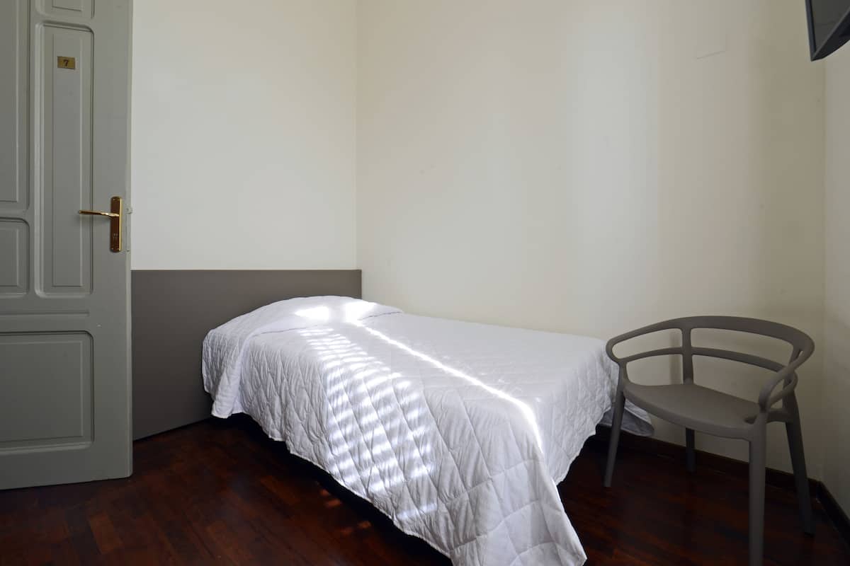 A small, single-bed room with a wooden floor, a white bedspread, a grey chair, and a white door with a brass handle.