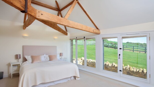 A bedroom with a large bed, a wooden ceiling beam, a nightstand, and a view of a green landscape through large windows. - Clayhanger Lodge  - Abbotsbury, Dorset (Abbotsbury)