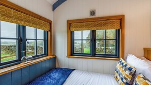 Une chambre à coucher avec un lit, deux fenêtres avec des rideaux, une vue sur la campagne et un mur de pierre à l’extérieur.