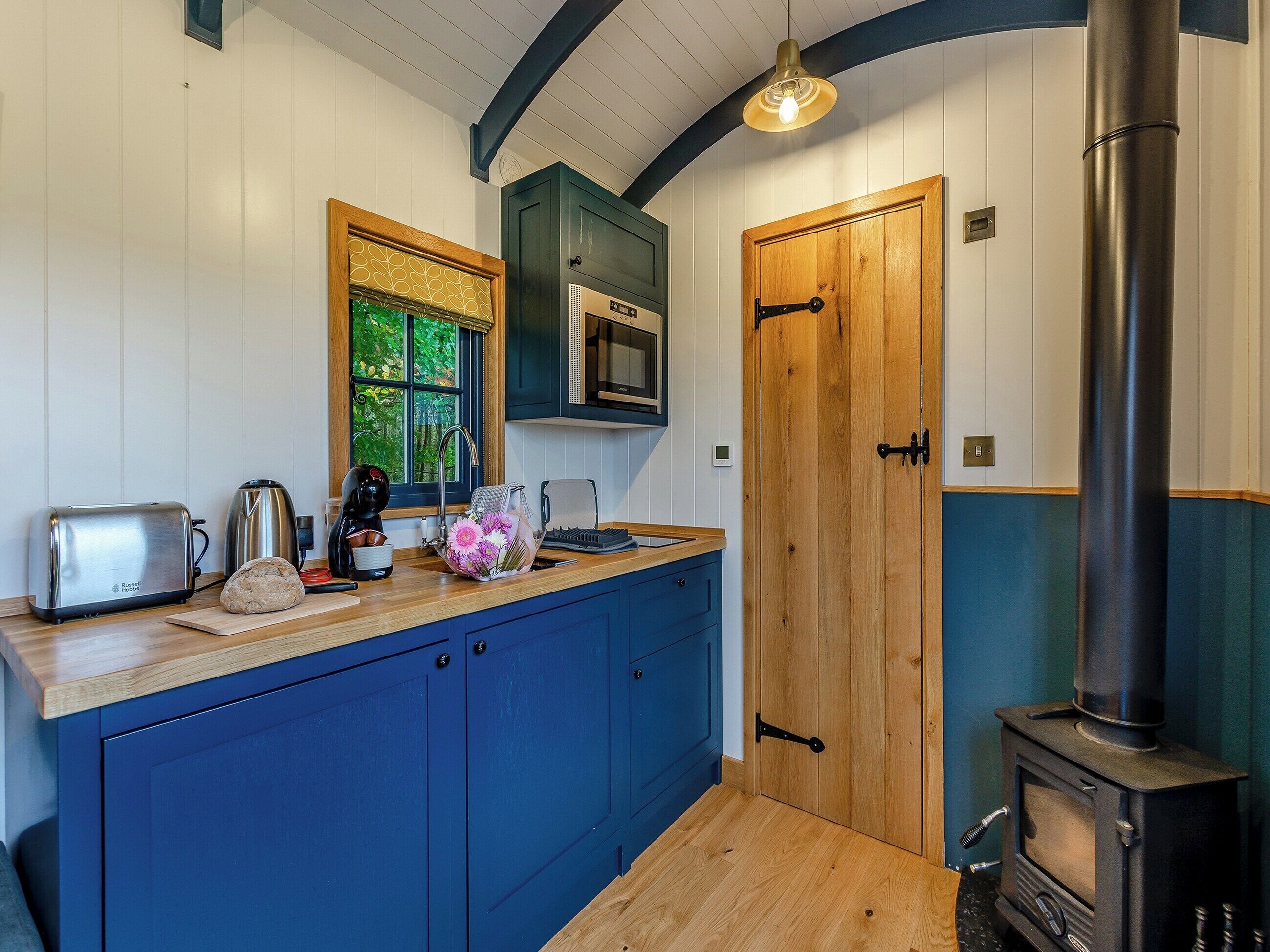 A compact kitchen with a toaster, kettle, and a microwave. There's a wooden door and a wood-burning stove.