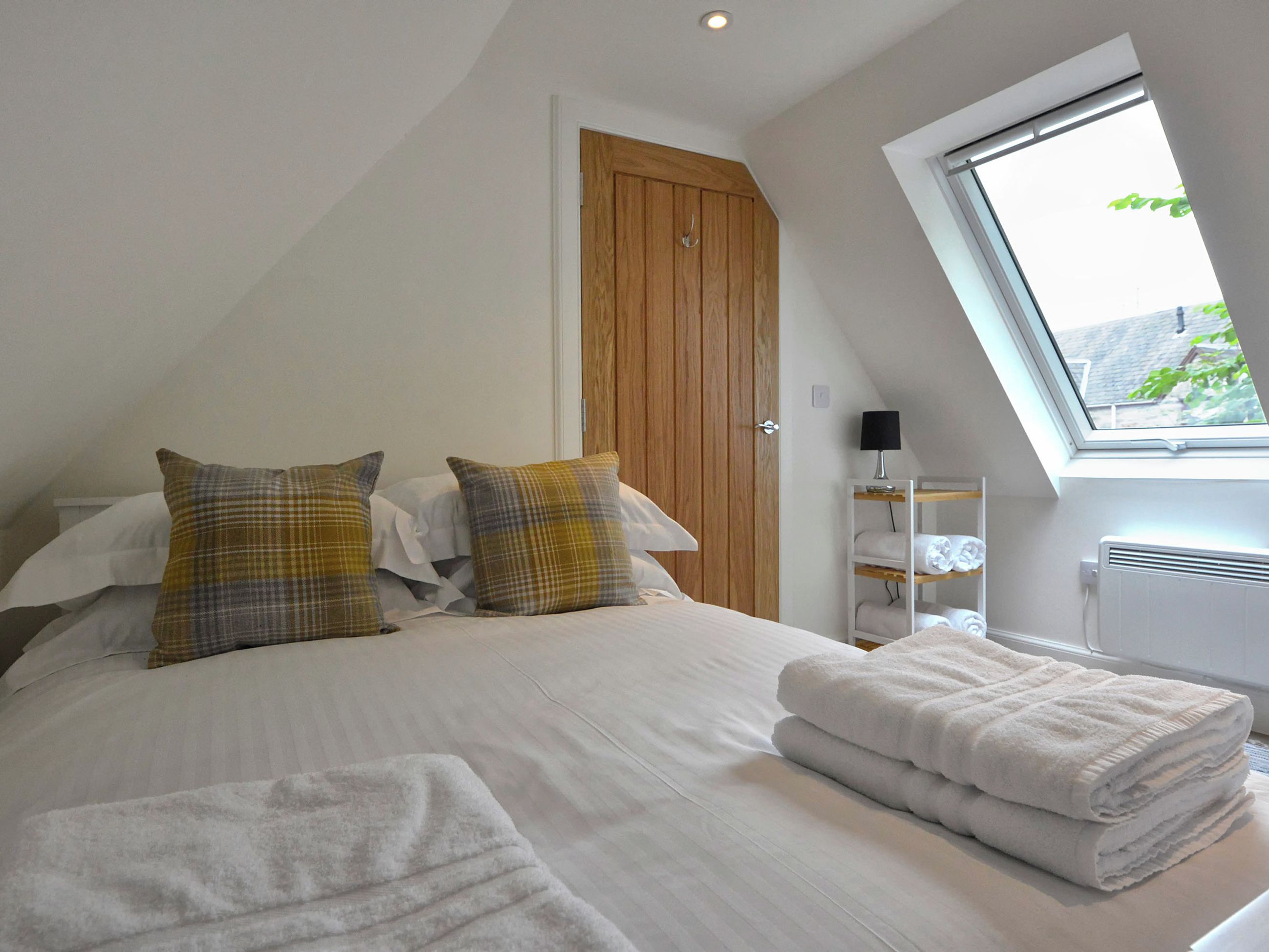 A bedroom with a bed, pillows, a wooden door, a window, and a shelf with towels.