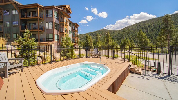 A wooden deck with a hot tub, outdoor seating, and a view of a multi-story building and forested mountains. - Clearwater Lofts Bldg 2 203 (Keystone)
