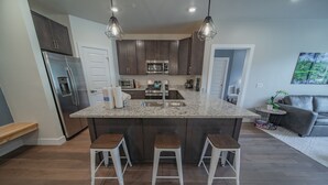 A modern kitchen with dark wood cabinets, stainless steel appliances, a granite countertop, and three bar stools. - Clearwater Lofts Bldg 2 203 (Keystone)