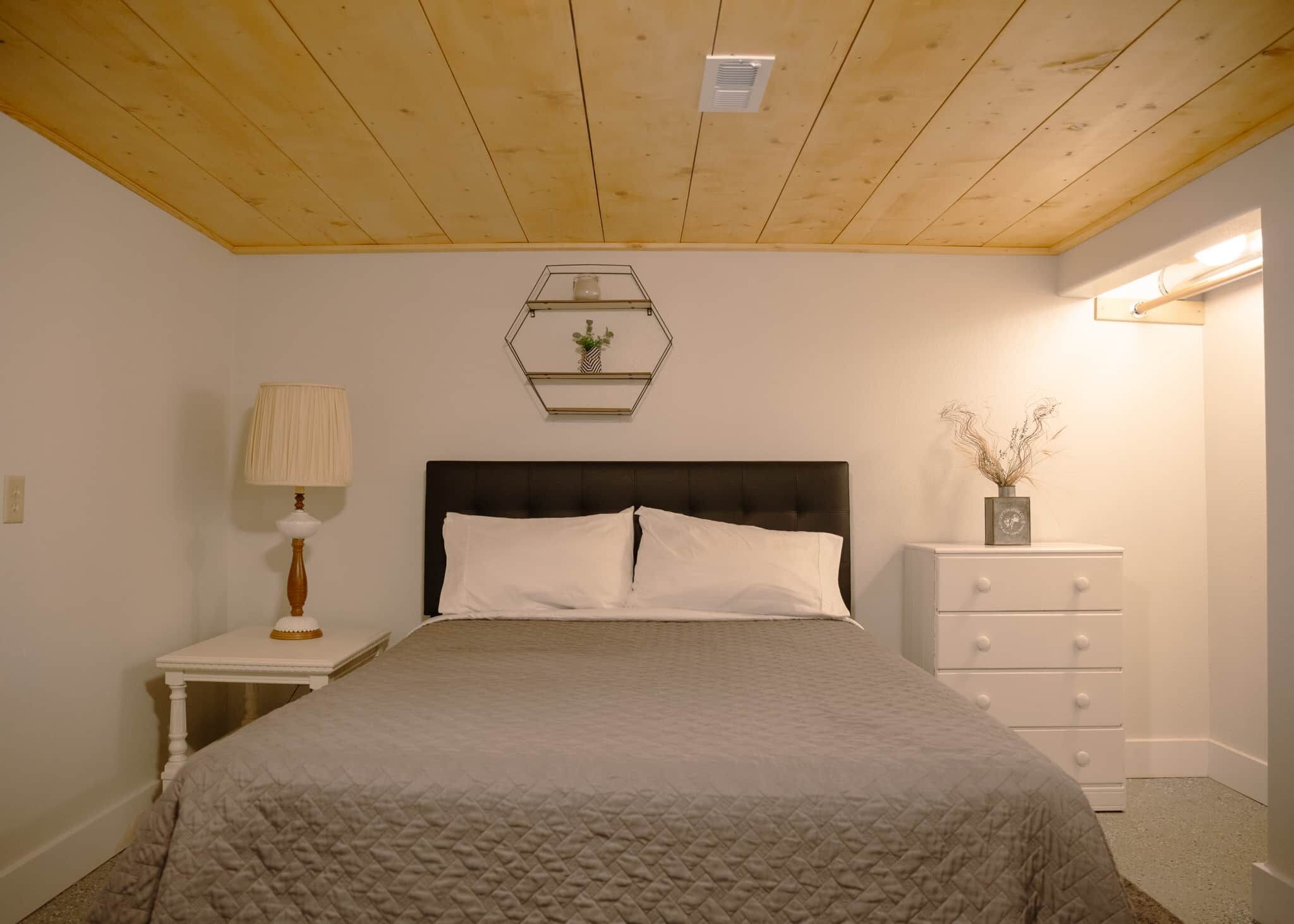 A bedroom with a bed, a nightstand, a dresser, a lamp, and a wall-mounted shelf with decorative items.