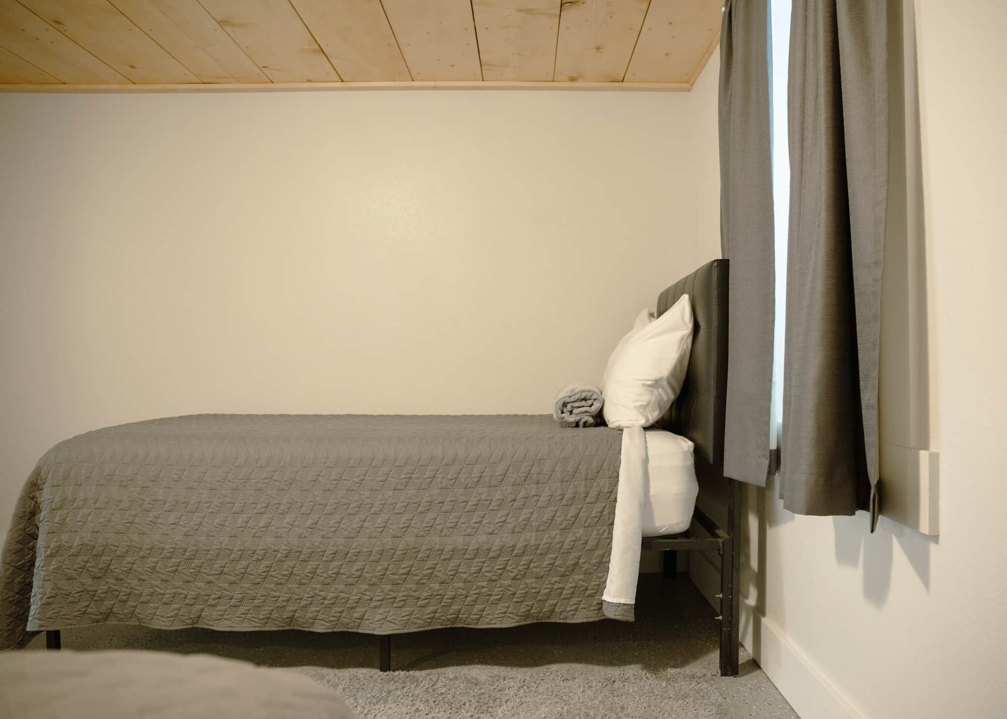 A single bed with a headboard, a pillow, and a blanket in a room with a wooden ceiling and a plain wall.