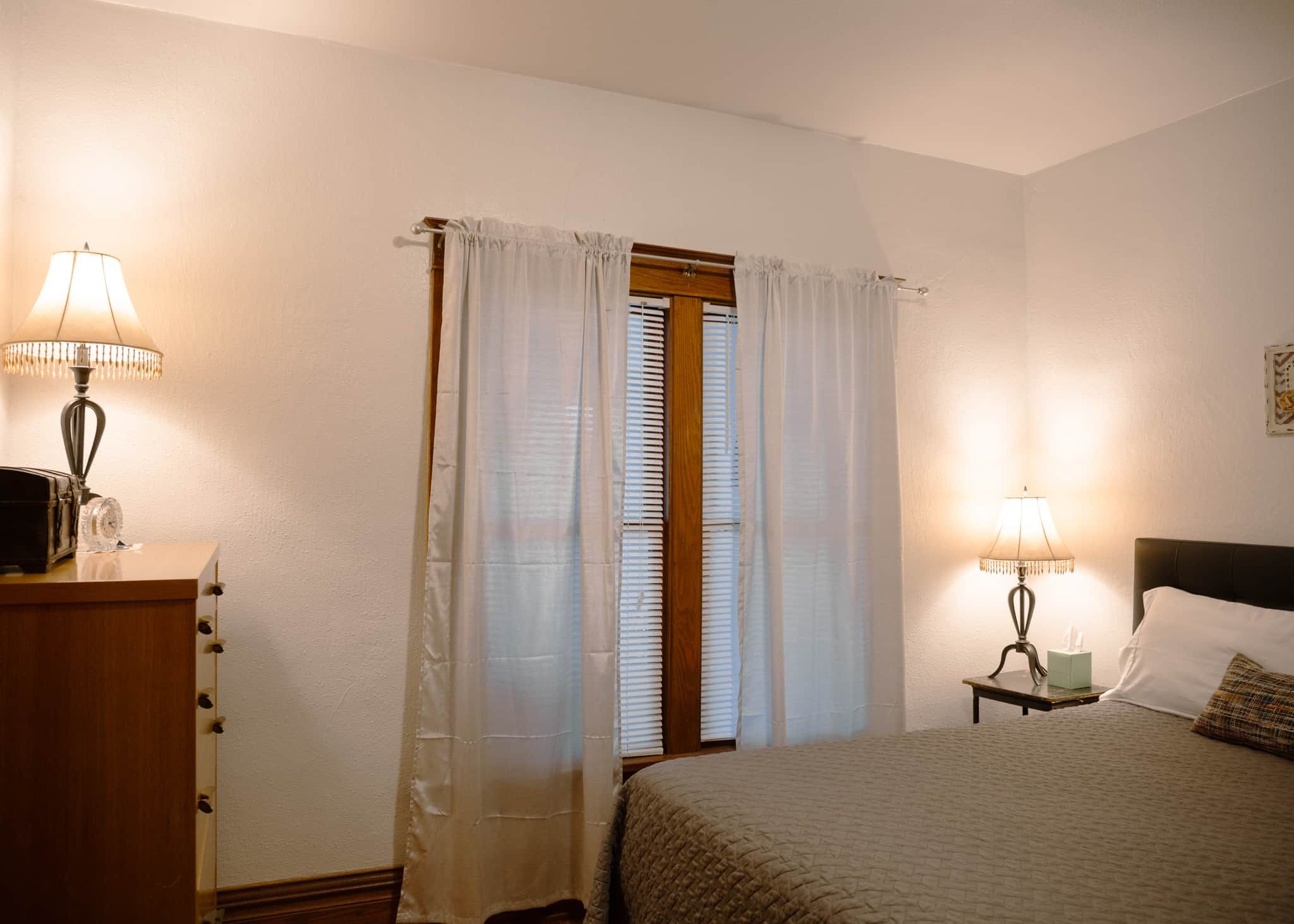 Un dormitorio con una cama, dos lámparas de noche, una mesita de noche, un aparador y una ventana con cortinas.