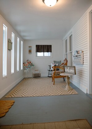 Une pièce avec un tapis à motifs chevrons, une chaise en bois portant une pancarte « Bienvenue », une petite table et une fenêtre avec un rideau foncé.