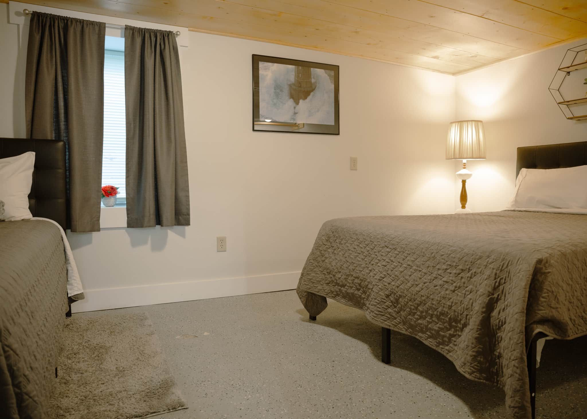 A room with two beds, a window with curtains, a wall-mounted lamp, and a painting.