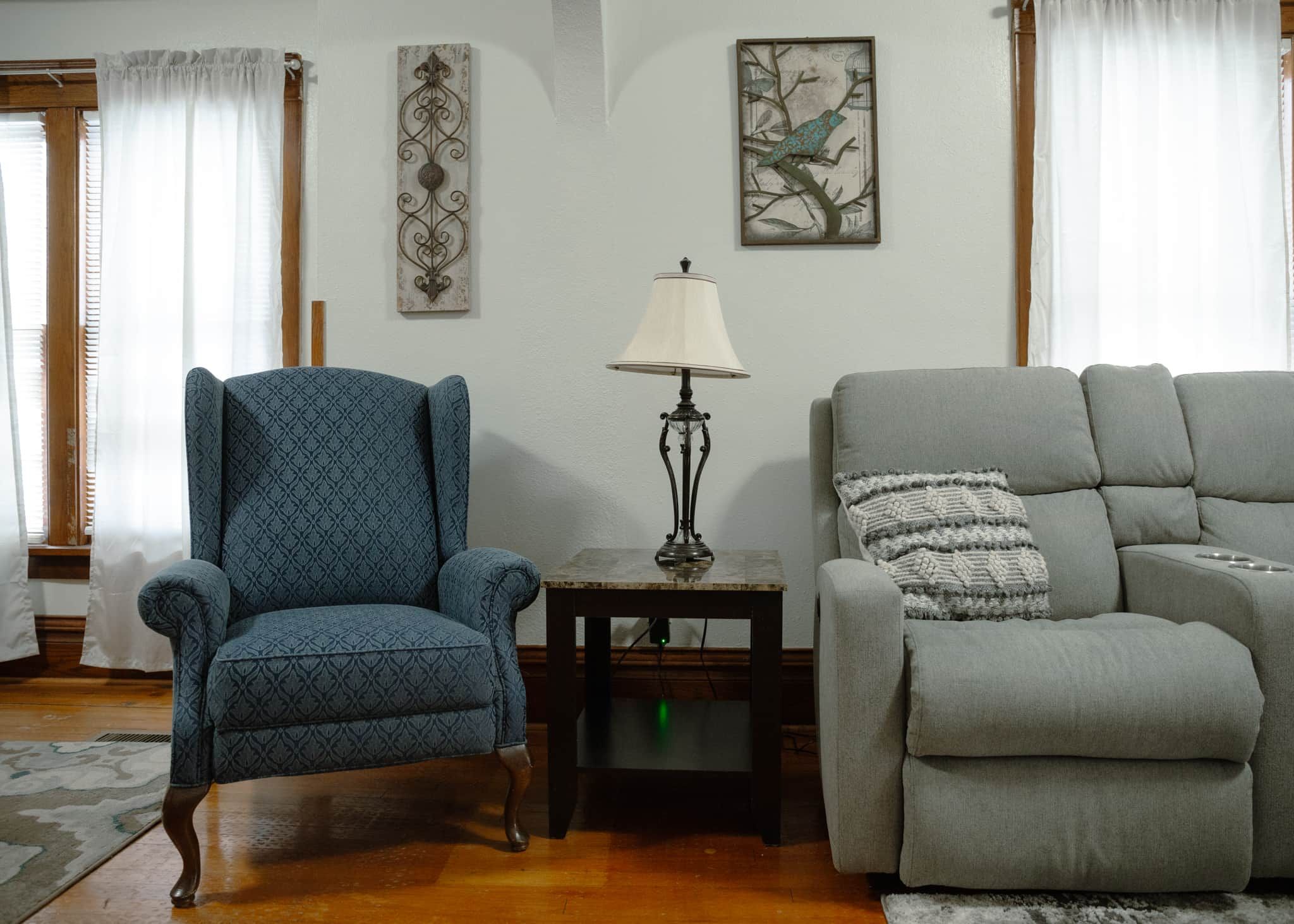 Una sala de estar con un sillón azul, un sofá gris, una mesa con una lámpara y dos adornos de pared.