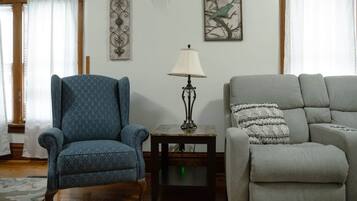 Una sala de estar con un sillón azul, un sofá gris, una mesa con una lámpara y dos adornos de pared.