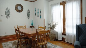 A dining area with a wooden table and chairs, a wall clock, decorative wall art, and a window with curtains.