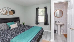 A bedroom with a bed, a bedside table, a window with curtains, a wall-mounted mirror, and a bathroom with a sink and toilet. - WinterBungalow (Myrtle Beach)
