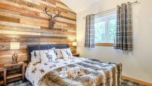 A bedroom with a wooden headboard, a deer-themed bedspread, a bedside table with a lamp, and a window with plaid curtains.