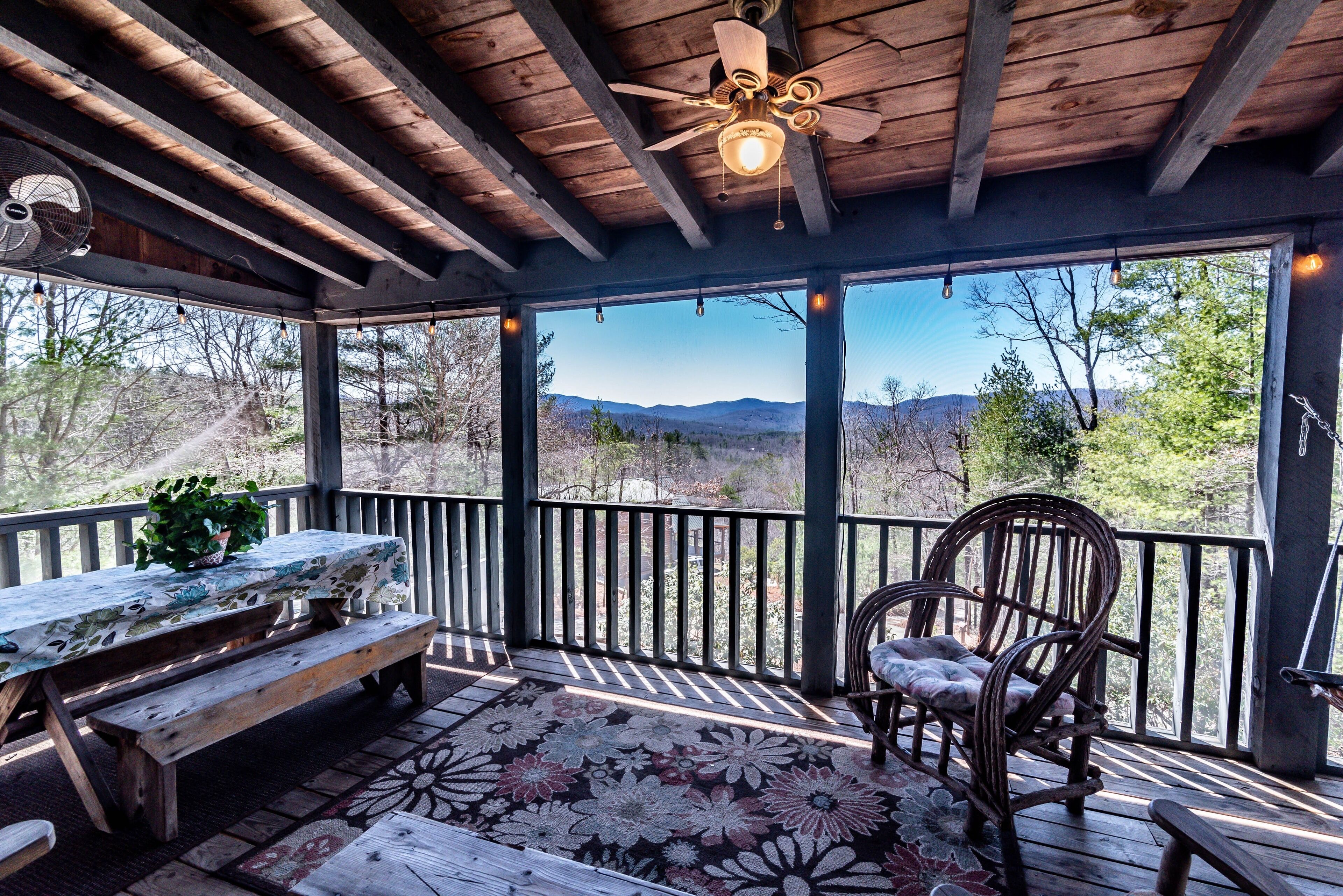 A covered deck with a wooden ceiling, a ceiling fan, a table with a floral tablecloth, a bench, and a chair.