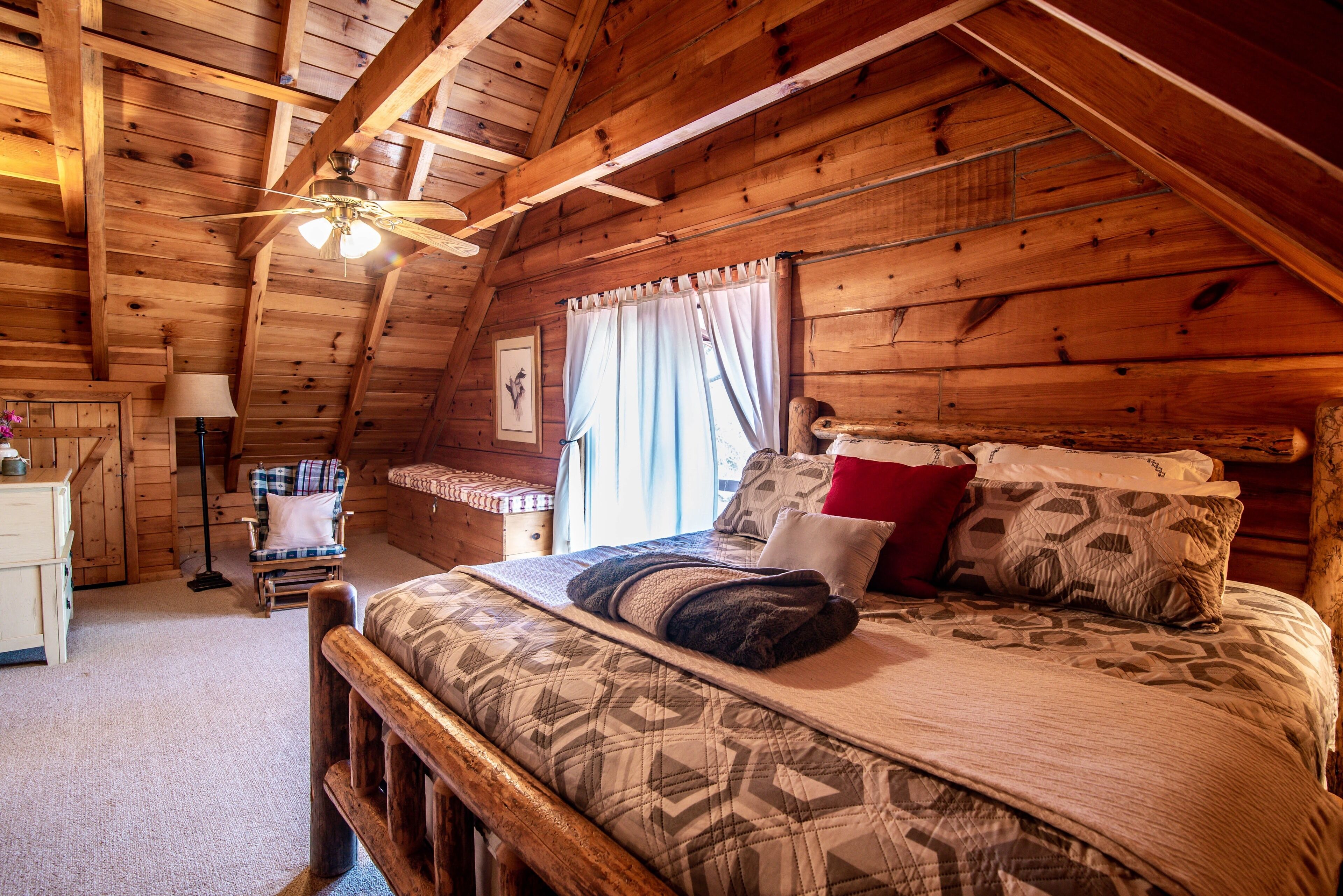 Quarto acolhedor de cabana de madeira, com teto de madeira, cama com roupa de cama estampada, uma cadeira, mesa de cabeceira e janela com cortinas.