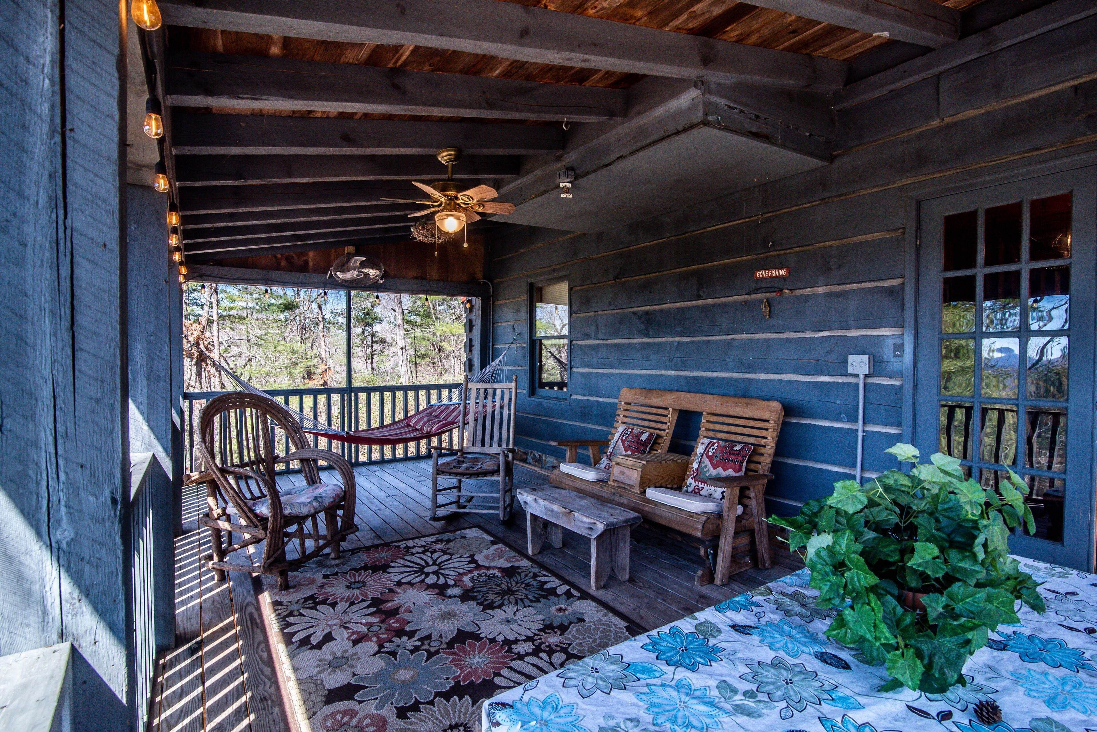 A wooden porch with a ceiling fan, rocking chairs, a bench, and a table with a floral tablecloth.
