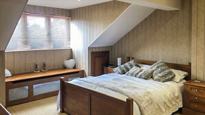 Une chambre avec un plafond en pente, un lit en bois, une table de chevet, un banc et une fenêtre avec des stores.