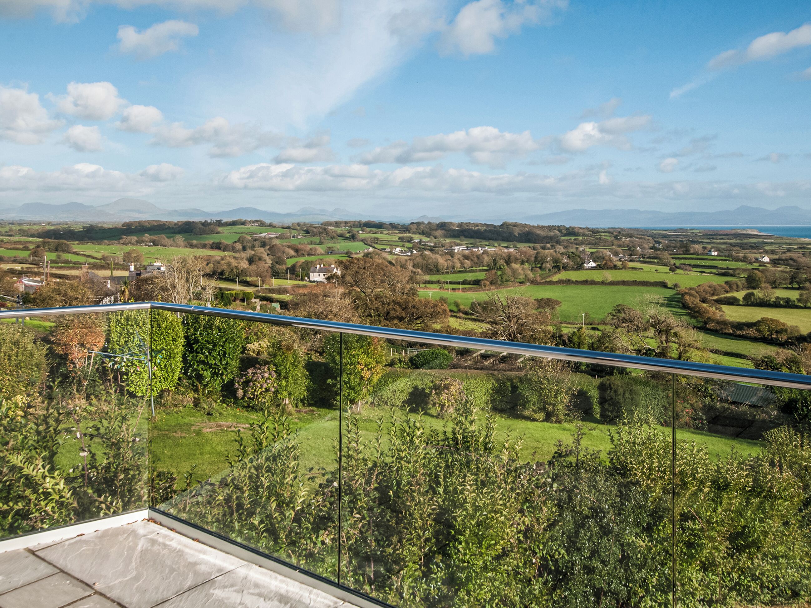A balcony with a glass railing overlooking a rural landscape with fields, trees, and distant mountains.
