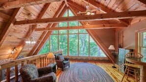 A cozy attic room with wooden beams, a large window, a round rug, and a seating area with a chair and a table. -  “The Creel” Luxury 2BR Cabin with loft & view (Townsend)