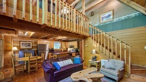 A rustic cabin interior with a wooden staircase, a dining area with a wooden table and chairs, a leather sofa, and a kitchen area. - “The Creel” Luxury 2BR Cabin with loft & view (Townsend)