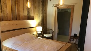 Una cama de madera con colchón blanco en una habitación con paredes y suelo de madera. Hay una mesita con una lámpara y una silla.