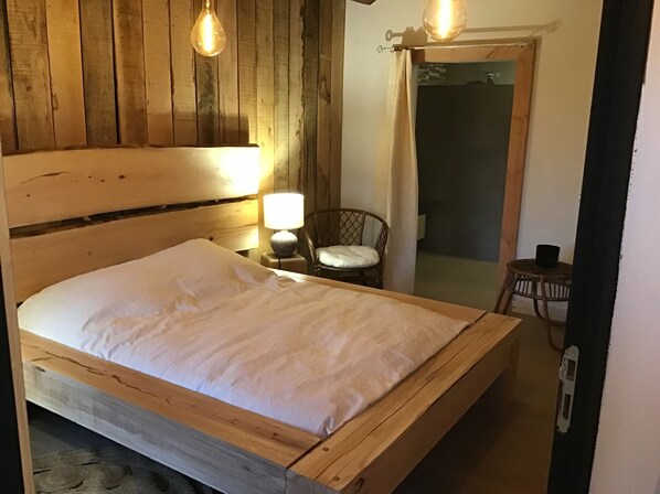 A wooden bed with a white mattress in a room with wooden walls and a wooden floor. There is a small table with a lamp and a chair. - Cottage of memories (Cézens)