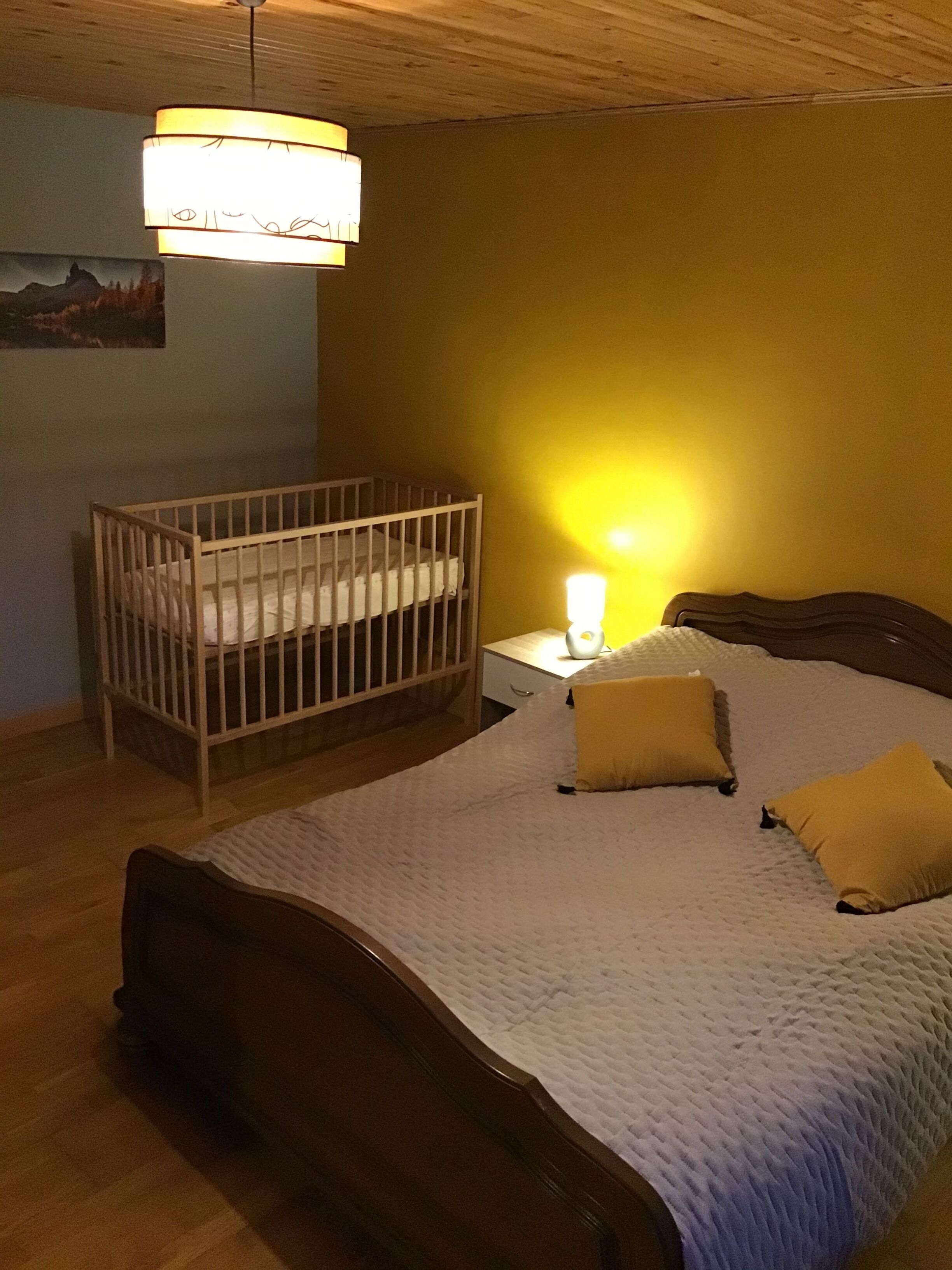 A bedroom with a wooden crib, a bed with a white quilt, and a bedside lamp.