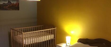 A bedroom with a wooden crib, a bed with a white quilt, and a bedside lamp.