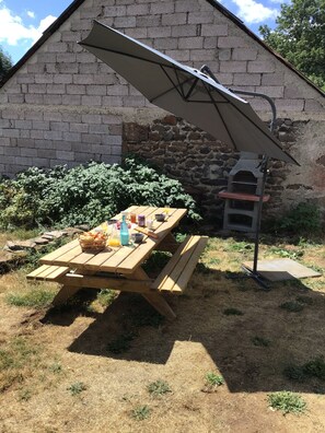 A wooden picnic table with a metal umbrella, a stone oven, and a brick wall in the background. - Cottage of memories (Cézens)