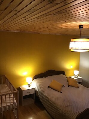 A bedroom with a wooden ceiling, a bed with a white quilt and two pillows, two bedside tables with lamps, and a small wooden crib. - Cottage of memories (Cézens)