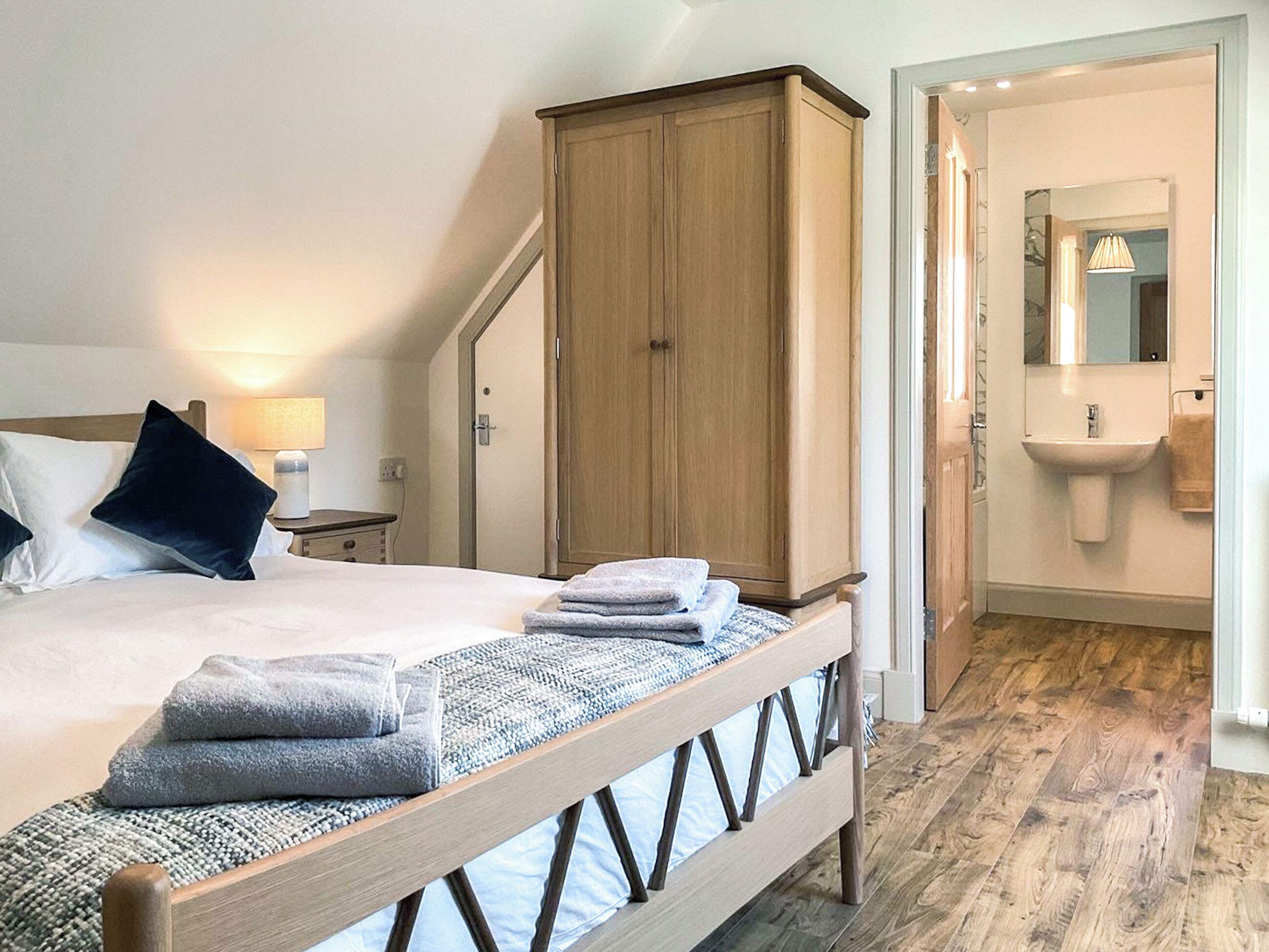 Een slaapkamer met een bed, een houten garderobe, een nachtkastje met een lamp, en een badkamer met een wastafel en spiegel.