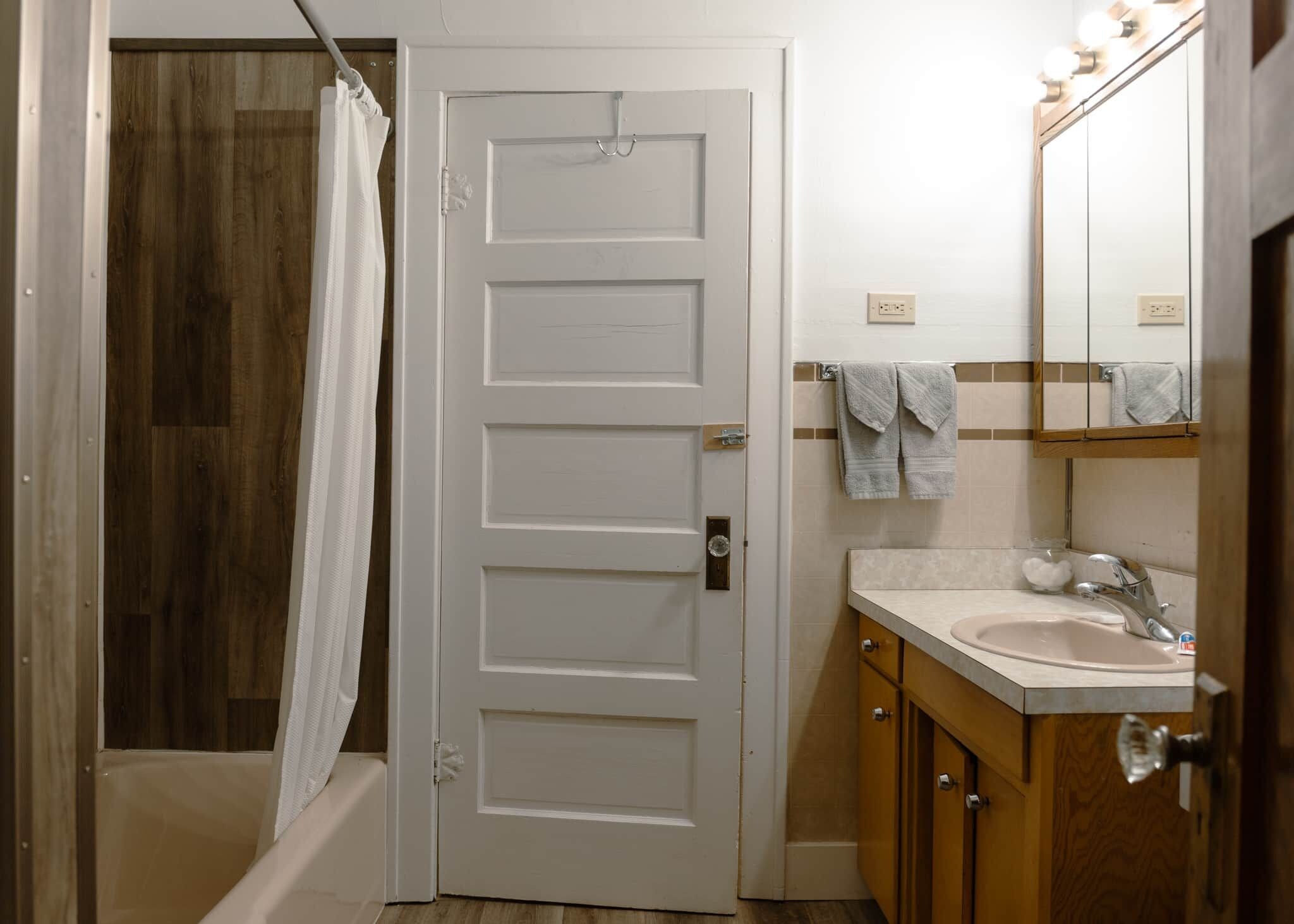 A bathroom with a white door, a shower curtain, a sink with a mirror, and towels on the wall.