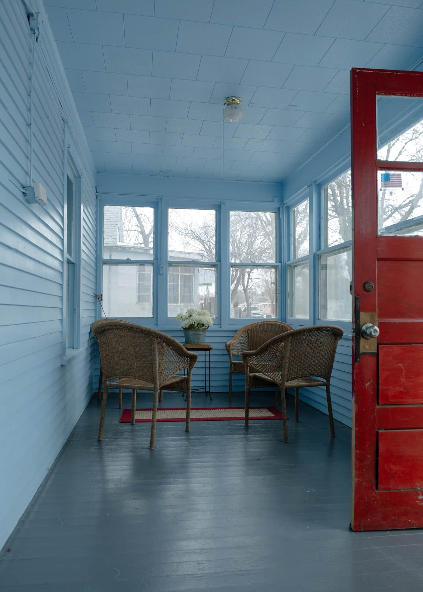 Una habitación con una puerta roja, dos sillas de mimbre, una mesita con un florero y ventanas amplias.