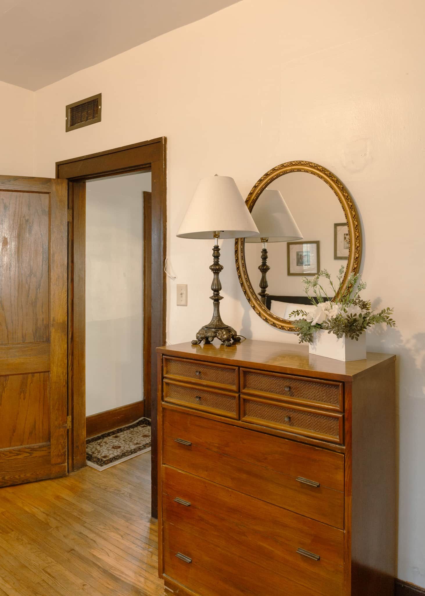 Un prestatge de fusta amb calaixos, una làmpada i un mirall amb marc daurat en una habitació amb una porta de fusta i una petita obertura a la paret.