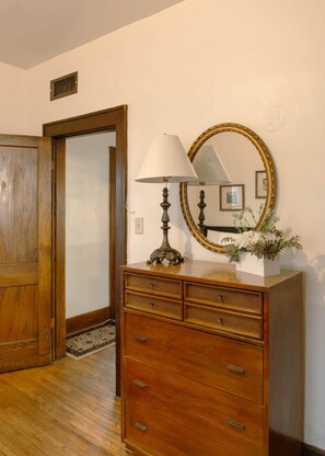 A wooden dresser with drawers, a lamp, and a mirror with a gold frame in a room with a wooden door and a small vent on the wall. - Red Roof Cottage – 2BR Mobridge Stay (Mobridge)
