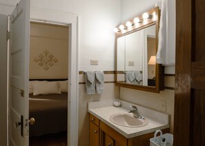 Casa de banho de hotel com lavatório, espelho e uma cama visível através de uma porta aberta.
