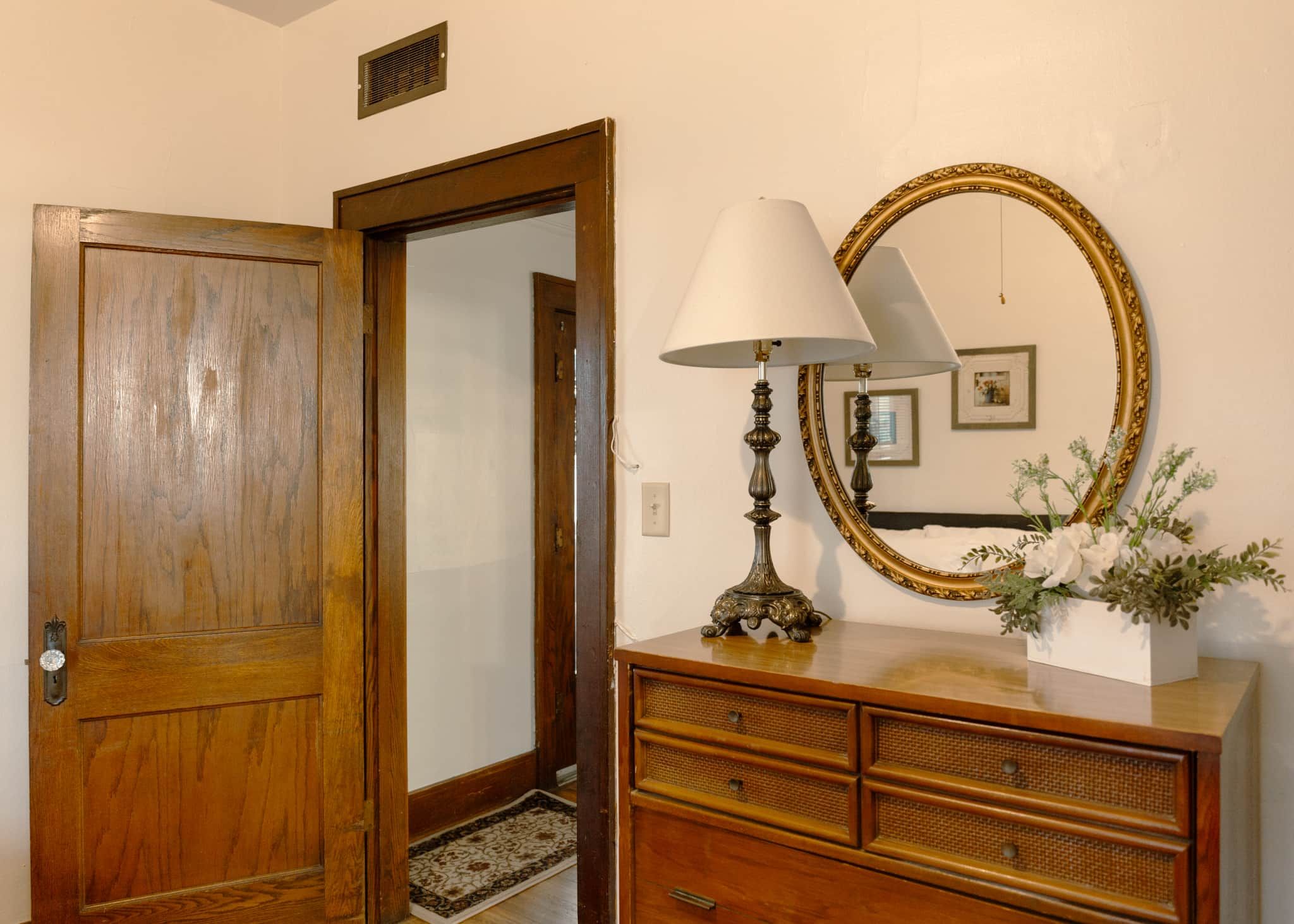 A wooden door, a lamp with a white shade, a mirror with a gold frame, and a dresser with a vase of flowers.