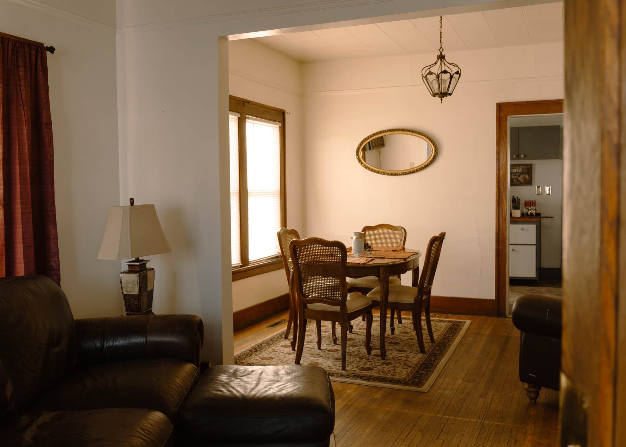 A living room with a brown leather sofa, a dining table with four chairs, a lamp, and a mirror on the wall.