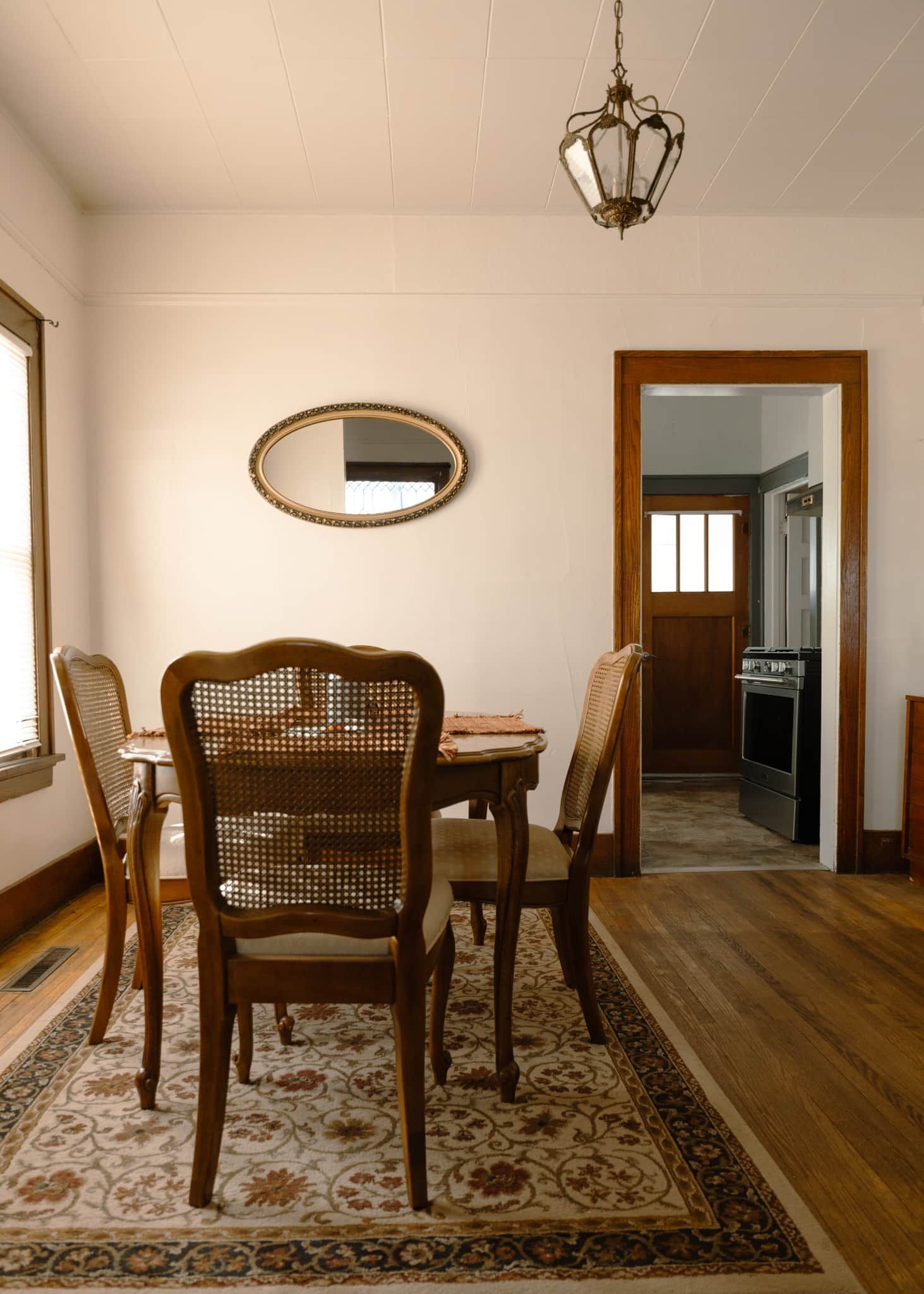 Un coin repas avec une table en bois, quatre chaises en osier, un tapis à motifs, un miroir au mur et un luminaire suspendu.