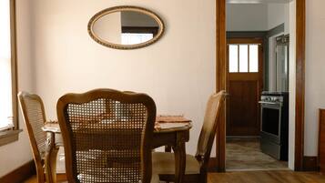 Un coin repas avec une table en bois, quatre chaises en osier, un tapis à motifs, un miroir au mur et un luminaire suspendu.