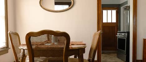 A dining area with a wooden table, four wicker chairs, a patterned rug, a mirror on the wall, and a hanging light fixture.