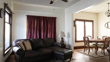 Una sala de estar con sofá de cuero, una mesa de comedor con sillas, un ventilador de techo y un ventanal con cortinas.