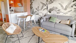 Una sala de estar moderna con un sofá gris, sillas blancas, una mesa de centro de madera y una cocina visible a través de una puerta abierta.