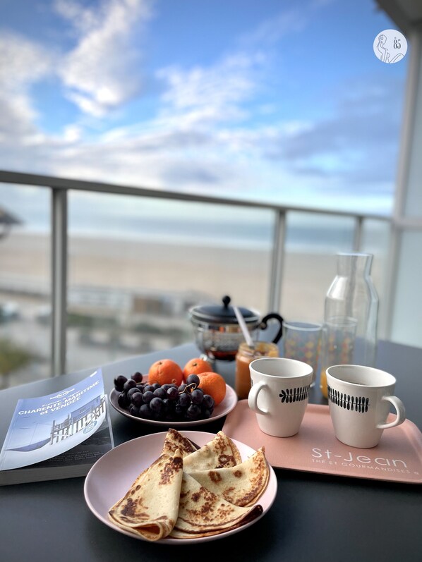 Un desayuno con vista al mar, que incluye una fuente de panqueques, un tazón de fruta y dos tazas de café sobre una bandeja.