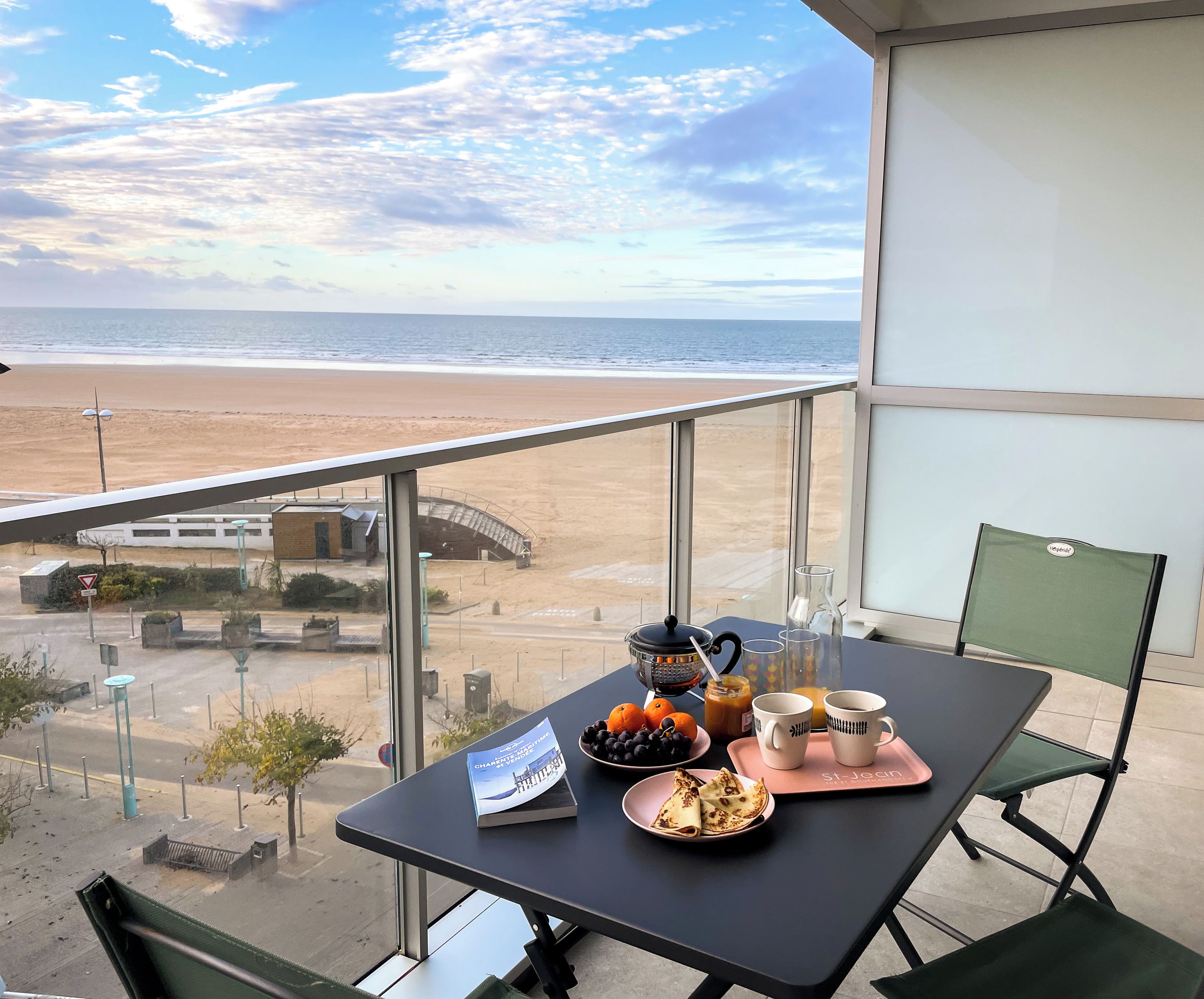 Un balcon avec une table préparée pour le petit-déjeuner, d’où l’on peut admirer une plage et un pont.