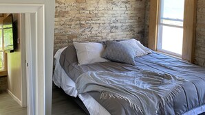 A bedroom with a brick wall, a ceiling fan, a bed with grey bedding, and a window with wooden frames. - Moments to Fiserv Forum and Downtown Milwaukee! (Milwaukee)