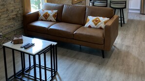 A modern living room with a brown leather sofa, a small table, and a kitchen in the background. - Moments to Fiserv Forum and Downtown Milwaukee! (Milwaukee)