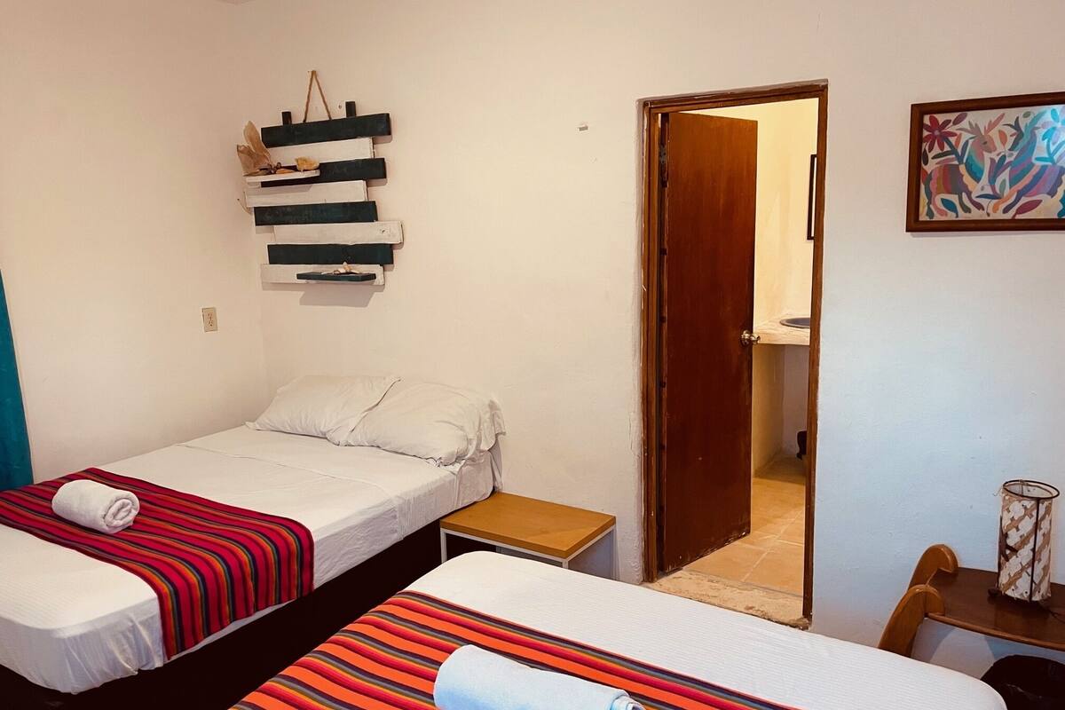 A small, clean room with two single beds, a wooden nightstand, a small table, and a door leading to another room.