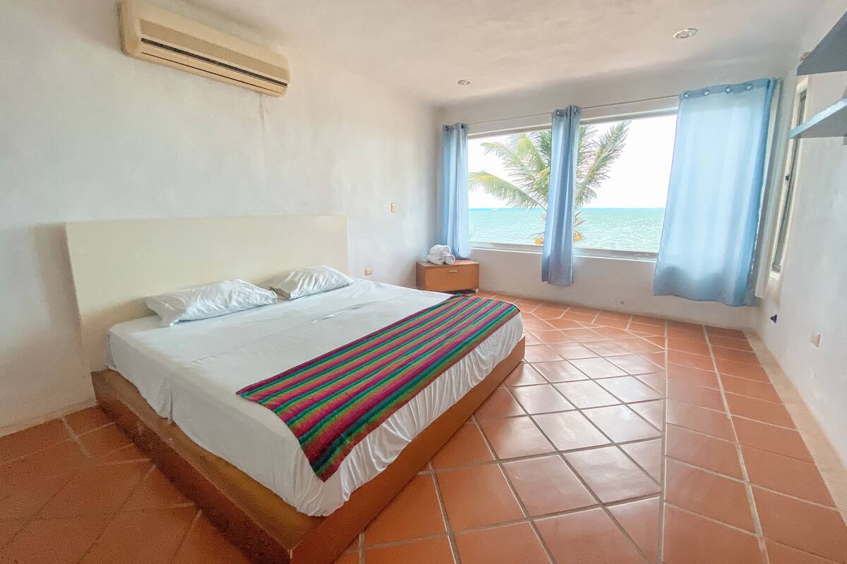 A bedroom with a bed, a nightstand, a window with curtains, and a view of the sea.