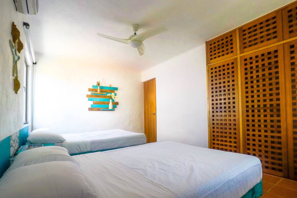 A room with two beds, a ceiling fan, a wooden door, and a decorative wall piece.